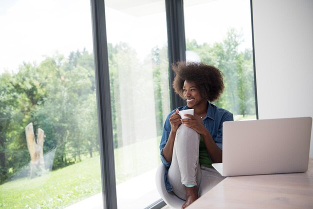 Young african american woman smiling sitting near bright window while looking at open laptop computer on table and holding white mug in her luxury home