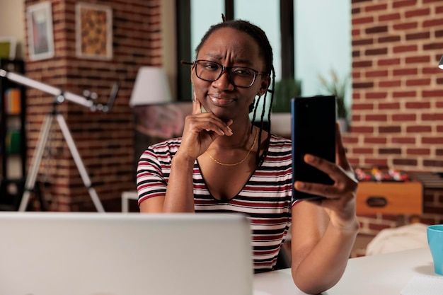 Молодая афроамериканка делает селфи на смартфоне, улыбаясь в переднюю камеру. Фрилансер делает портретное фото на мобильный телефон на рабочем месте в домашнем офисе, удаленный работник разговаривает по видеозвонку