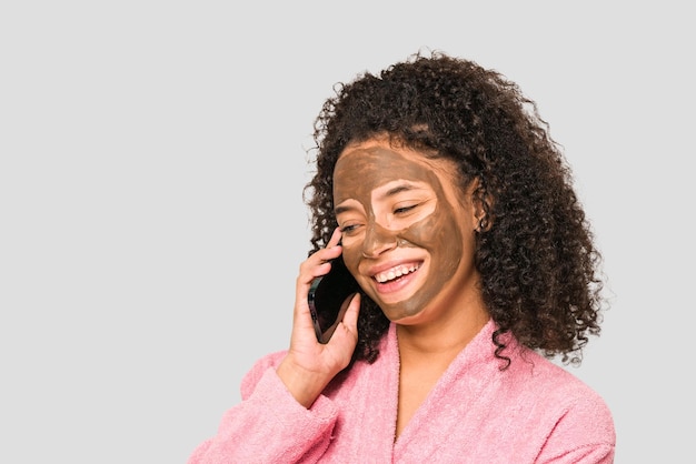 Young african american woman talking on phone while she is applying a facial mask isolated