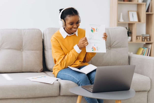 A young african-american woman talks on a video call and shows\
something on paper in the living room at home, the concept of\
distance learning from home.