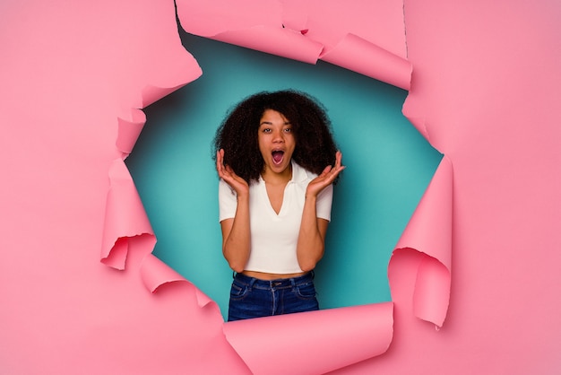 Giovane donna afro-americana in carta strappata isolato su sfondo blu sorpreso e scioccato.