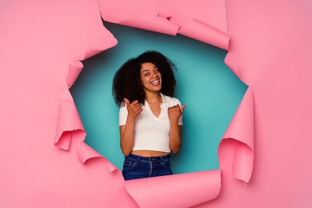 Young African American woman in torn paper isolated on blue raising both thumbs up, smiling and confident.