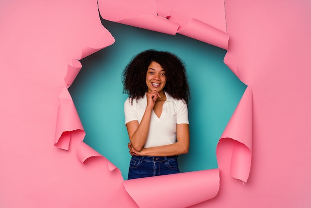 Young African American woman in torn paper isolated on blue smiling happy and confident, touching chin with hand.