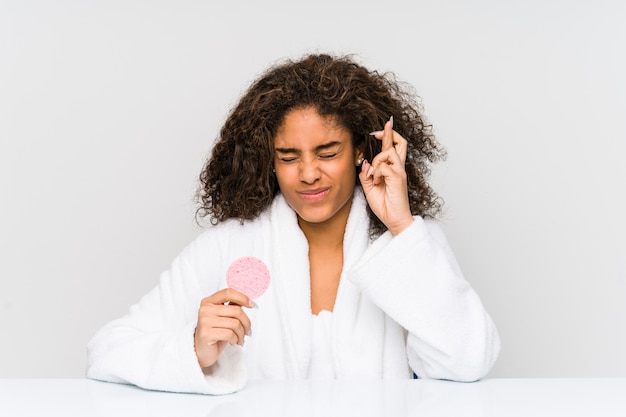 Young african american woman using a facial disk crossing fingers for having luck