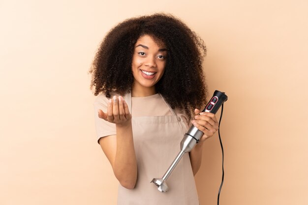 Young african american woman using hand blender on beige wall inviting to come