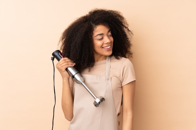 Young african american woman using hand blender on beige wall looking to the side