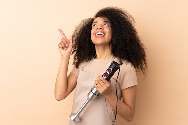 Photo young african american woman using hand blender on beige wall pointing with the index finger