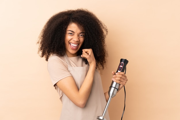 Young african american woman using hand blender isolated on beige making strong gesture