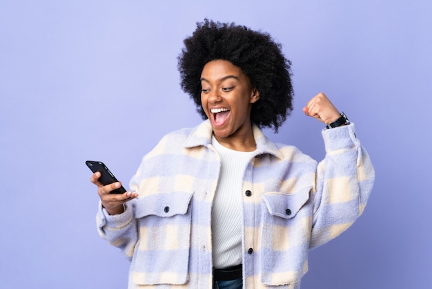 Young African American woman using mobile phone isolated on purple celebrating a victory