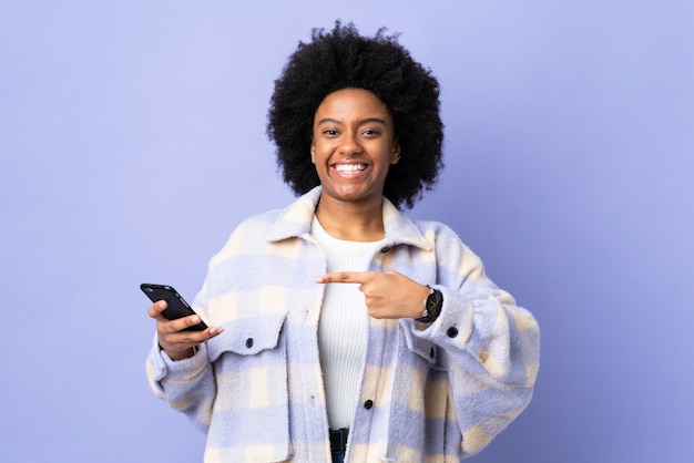 Young African American woman using mobile phone isolated on purple and pointing it