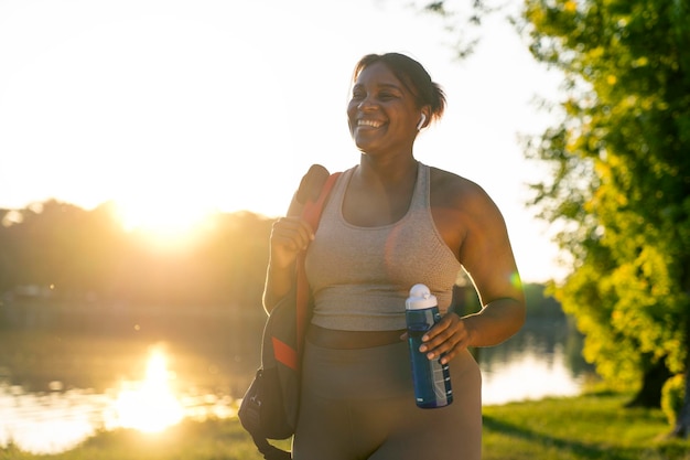 若いアフリカ系アメリカ人の女性が夏の日にバッグと水のボトルを持って公園を歩いています