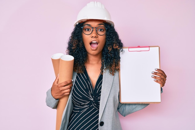 Young african american woman wearing architect hardhat holding blueprints and clipboard afraid and shocked with surprise and amazed expression, fear and excited face.