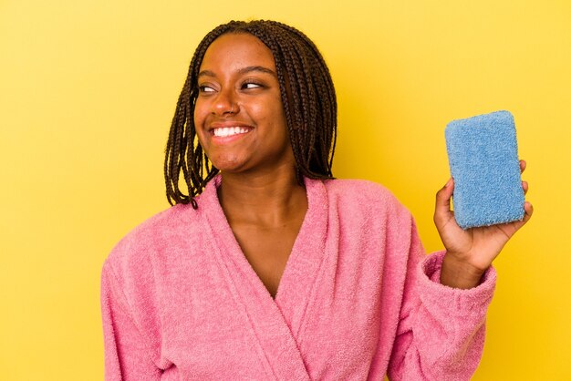 노란색 배경에 격리된 파란색 스폰지를 들고 목욕 가운을 입은 젊은 아프리카계 미국인 여성은 옆으로 웃고, 명랑하고 유쾌해 보입니다.