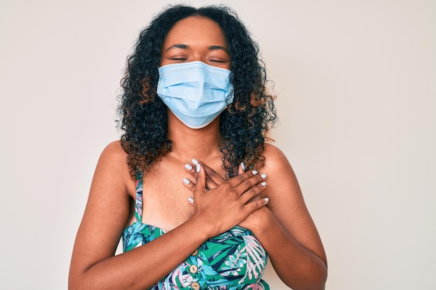 Giovane donna afroamericana che indossa una maschera medica sorridente con le mani sul petto, gli occhi chiusi con un gesto grato sul viso. concetto di salute.