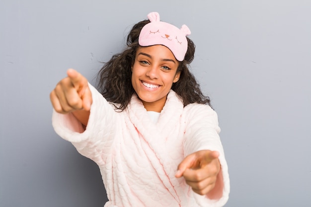 Young african american woman wearing a pajamas and a sleep mask cheerful smiles pointing to front.