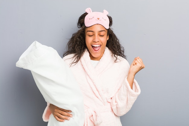 Young african american woman wearing a pajamas and a sleep mask holding a pillow cheering carefree and excited. Victory concept.