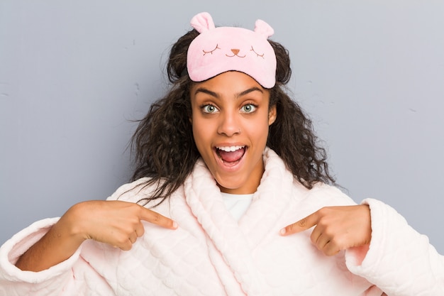 Young african american woman wearing a pajamas and a sleep mask surprised pointing at herself