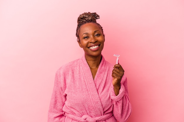 young african american woman wearing pink bathrobe holding razor blade isolated on pink background happy, smiling and cheerful.