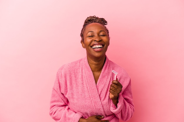 young african american woman wearing pink bathrobe holding razor blade isolated on pink background laughing and having fun.