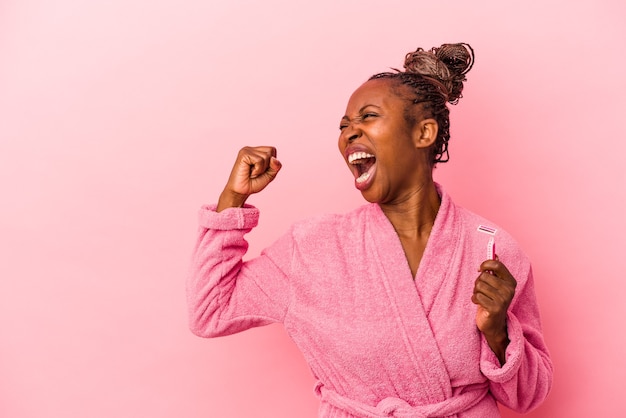 Giovane donna afroamericana che indossa accappatoio rosa tenendo la lama di rasoio isolata su sfondo rosa alzando il pugno dopo una vittoria, concetto vincitore.