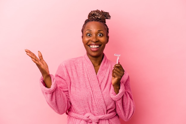 Молодая афро-американская женщина в розовом халате, держащая бритвенное лезвие, изолированное на розовом фоне, получает приятный сюрприз, взволнована и поднимает руки.