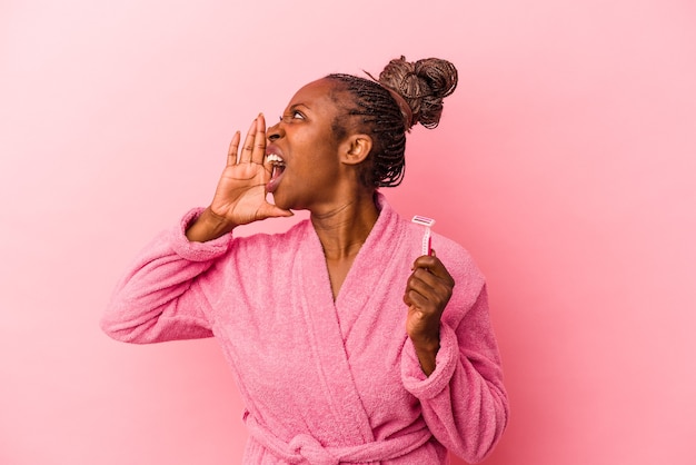 Young african american woman wearing pink bathrobe holding razor blade isolated on pink background shouting and holding palm near opened mouth.