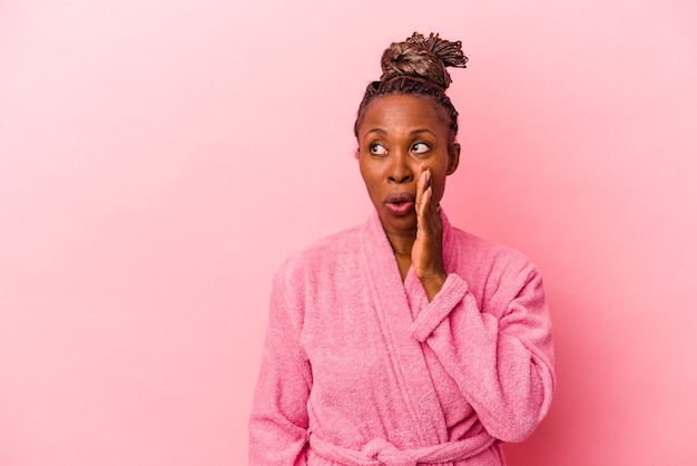 Young african american woman wearing pink bathrobe isolated on\
pink background is saying a secret hot braking news and looking\
aside