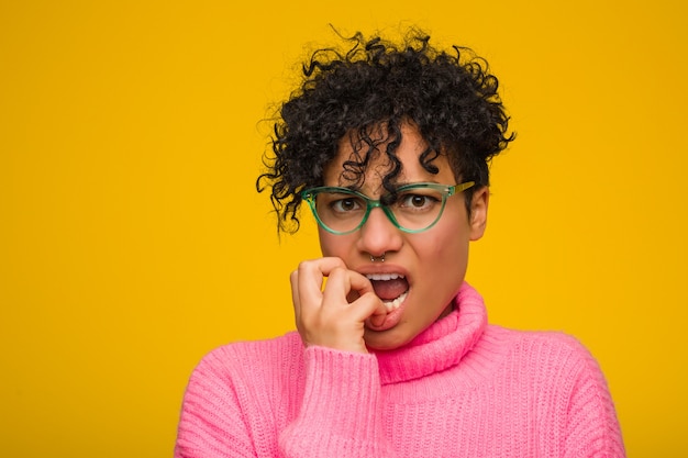 Young african american woman wearing a pink sweater biting fingernails, nervous and very anxious.