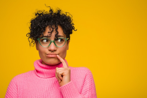 Giovane donna afroamericana che indossa un maglione rosa che guarda lateralmente con espressione dubbiosa e scettica.