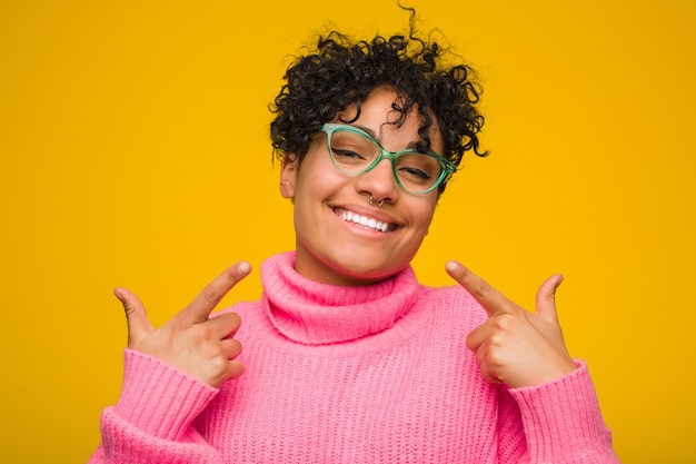 Young african american woman wearing a pink sweater smiles, pointing fingers at mouth.