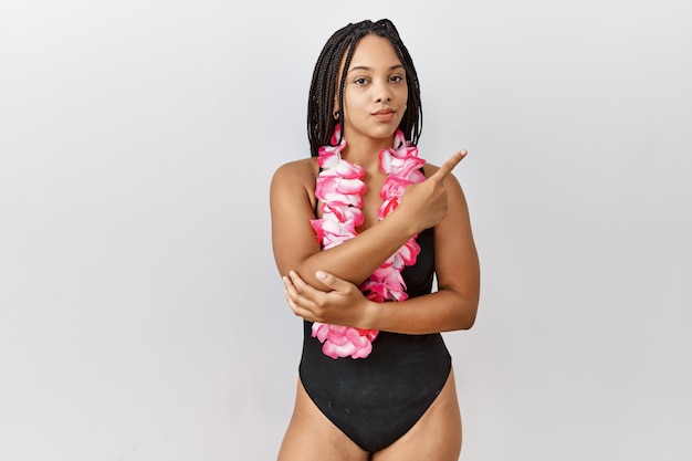 Young african american woman wearing swimsuit and hawaiian lei pointing with hand finger to the side showing advertisement, serious and calm face