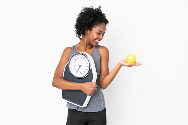 Young African American woman on white wall holding a weighing machine while looking an apple