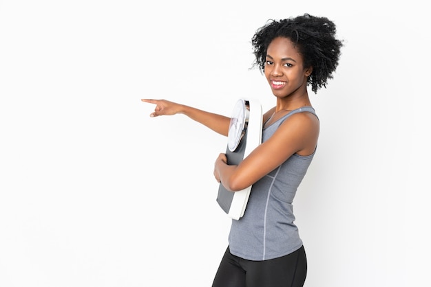 Young African American woman on white wall with weighing machine and pointing side
