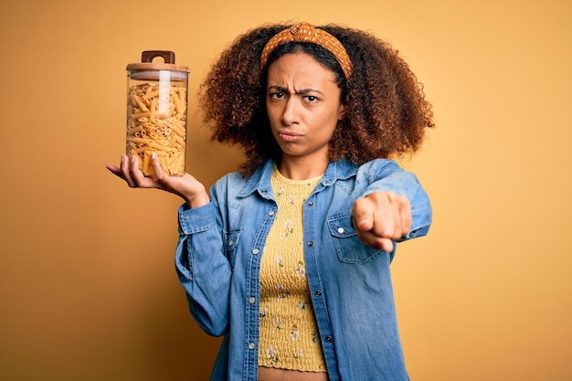 Young african american woman with afro hair holding jar with healthy macaroni pasta pointing with finger to the camera and to you hand sign positive and confident gesture from the front