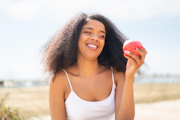 Молодая афроамериканка с яблоком на открытом воздухе с счастливым выражением лица