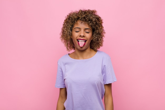 Young african american woman with cheerful, carefree, rebellious attitude, joking and sticking tongue out, having fun against pink wall