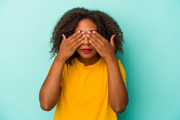 Young african american woman with curly hair isolated on blue background afraid covering eyes with hands.