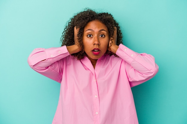 Молодая афро-американская женщина с вьющимися волосами изолирована на синем фоне, закрывая уши руками, пытаясь не слышать слишком громкий звук.