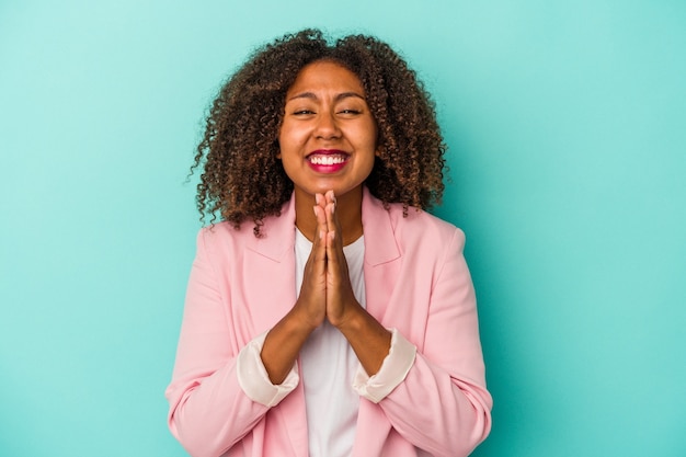 Young african american woman with curly hair isolated on blue background holding hands in pray near mouth, feels confident.