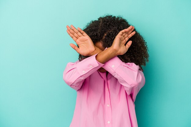 Young african american woman with curly hair isolated on blue background keeping two arms crossed, denial concept.