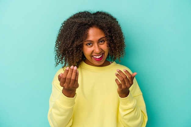 Young african american woman with curly hair isolated on blue background pointing with finger at you as if inviting come closer.