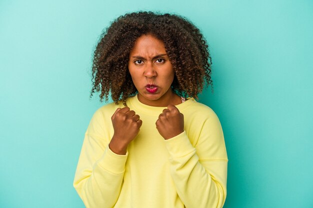 Young african american woman with curly hair isolated on blue background showing fist to camera, aggressive facial expression.