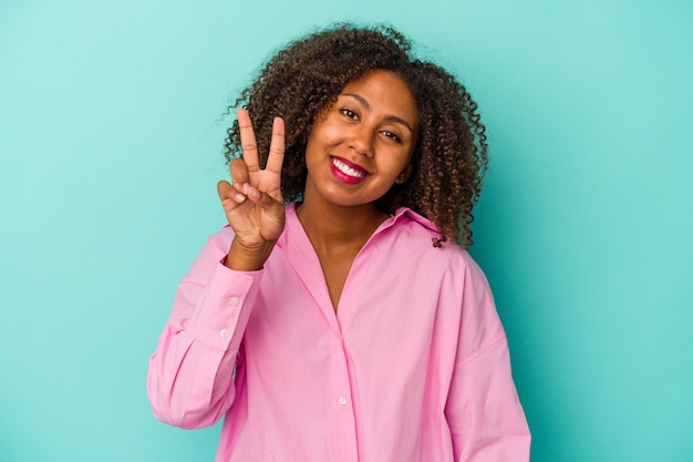 Молодая афро-американская женщина с вьющимися волосами, изолированными на синем фоне, показывая номер два пальцами.