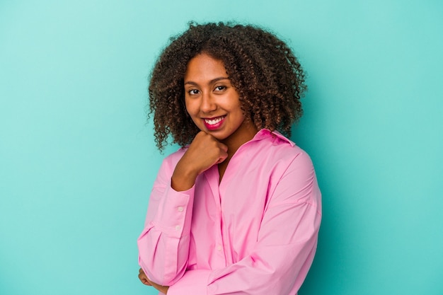 Young african american woman with curly hair isolated on blue background smiling happy and confident, touching chin with hand.