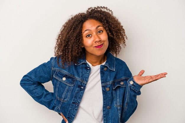 Young african american woman with curly hair isolated on white background showing a copy space on a palm and holding another hand on waist.