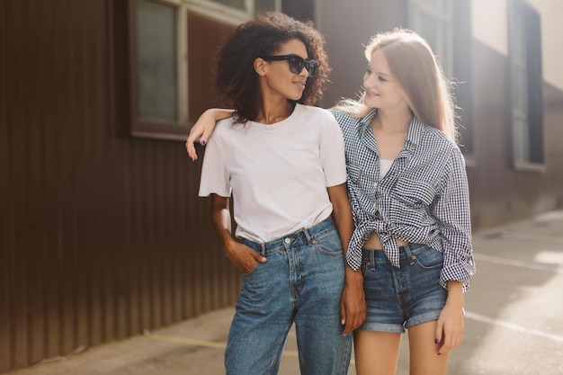 Young african american woman with dark curly hair in sunglasses and Tshirt and pretty woman with blond hair in shirt dreamily looking at each other while spending time together