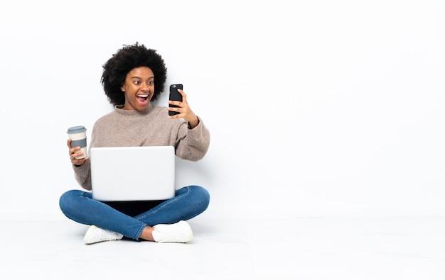 Молодая афро-американских женщина с ноутбуком, сидя на полу, держа кофе на вынос и мобильный
