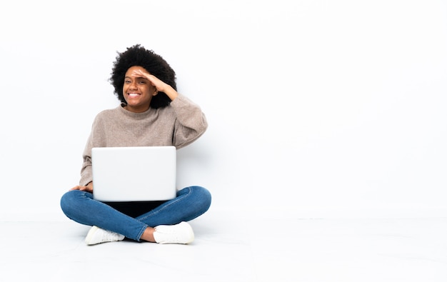 Young African American woman with a laptop sitting on the floor looking far away with hand to look something