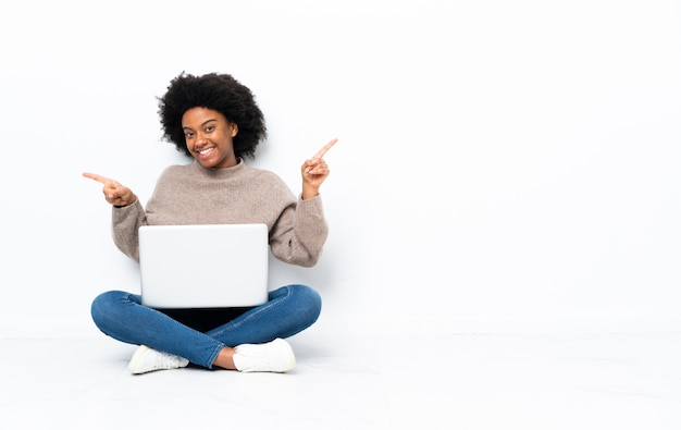 Giovane donna afroamericana con un computer portatile che si siede sul pavimento che indica il dito ai laterali e felice