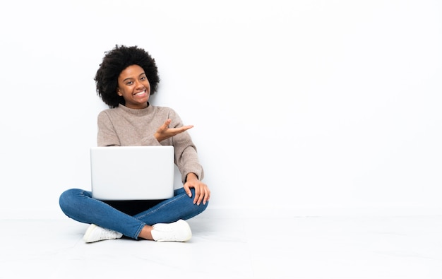 Young African American woman with a laptop sitting on the floor presenting an idea while looking smiling towards